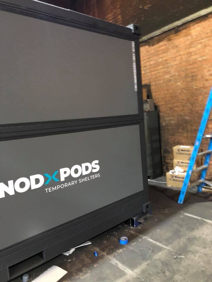 Gray and black NodPod shelter in a brick-walled room.  A blue ladder and boxes are next to it.