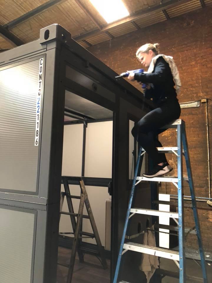 Woman on a ladder, using a tool to work on a dark-framed structure. Inside is a white interior. Industrial setting.