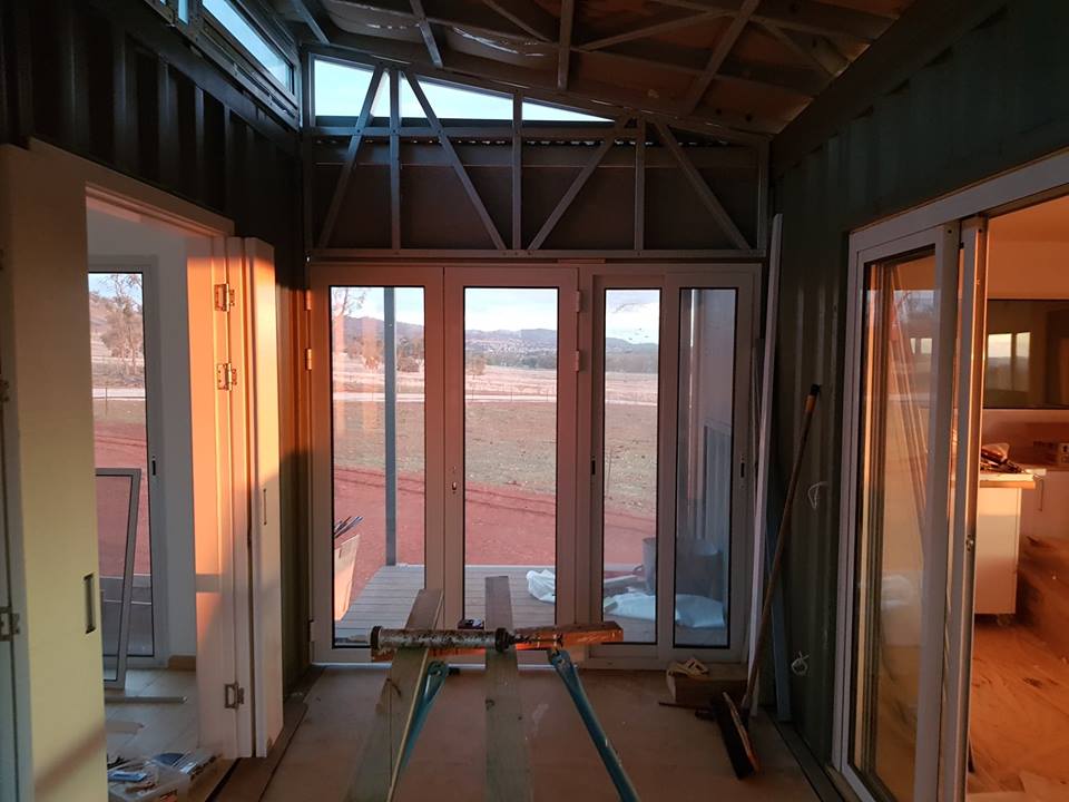 Interior of a room under construction with white framed windows and doors, metal framing, and a view of a field.