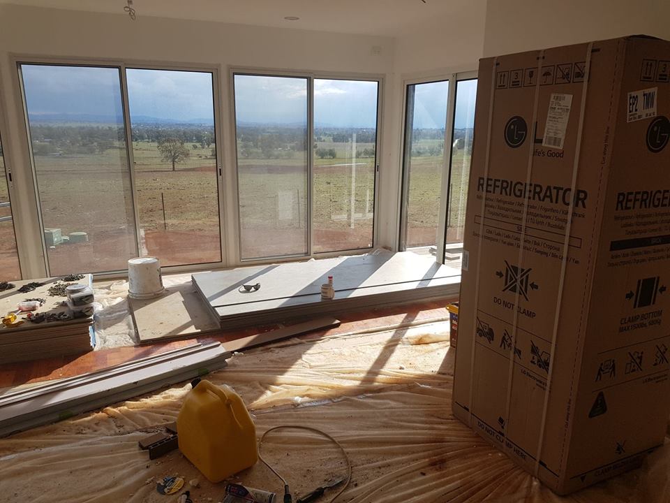 A new refrigerator in a cardboard box sits in a room with large windows overlooking a rural landscape.