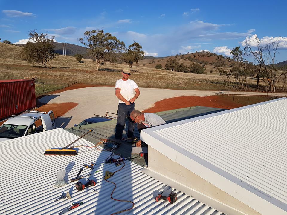 Two men working on a white corrugated metal roof outdoors, with tools and a van nearby, overlooking a rural landscape.