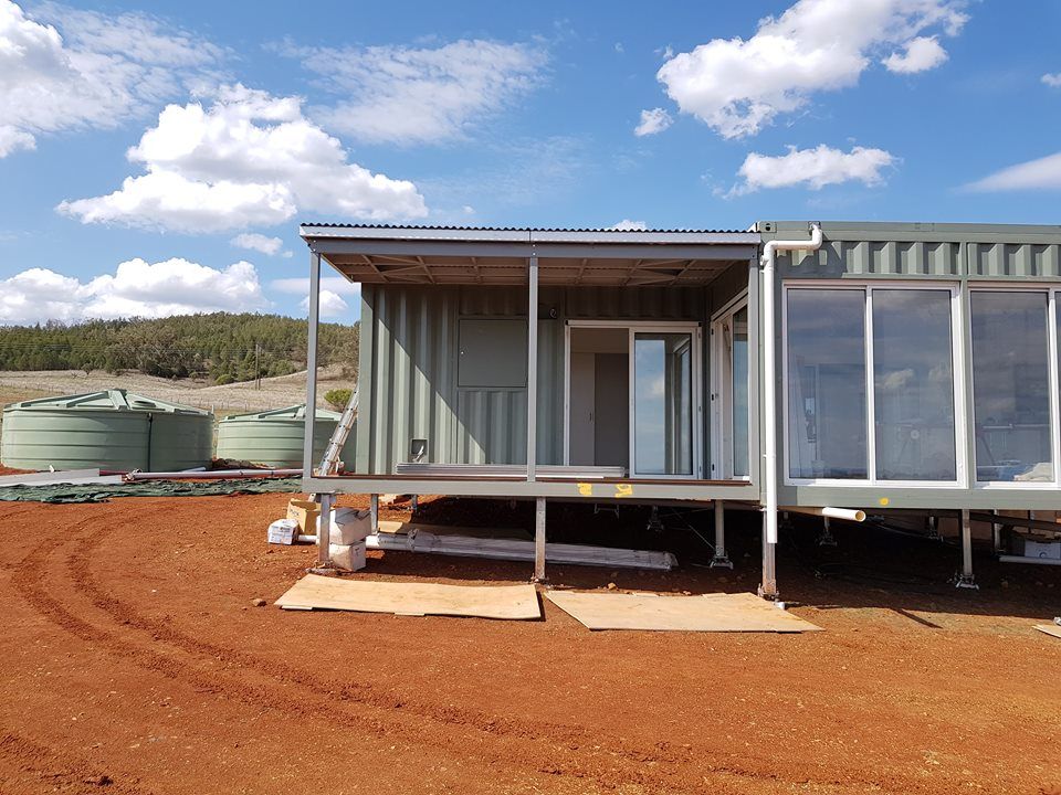 Shipping container home under construction on a red dirt lot, with water tanks in the background.