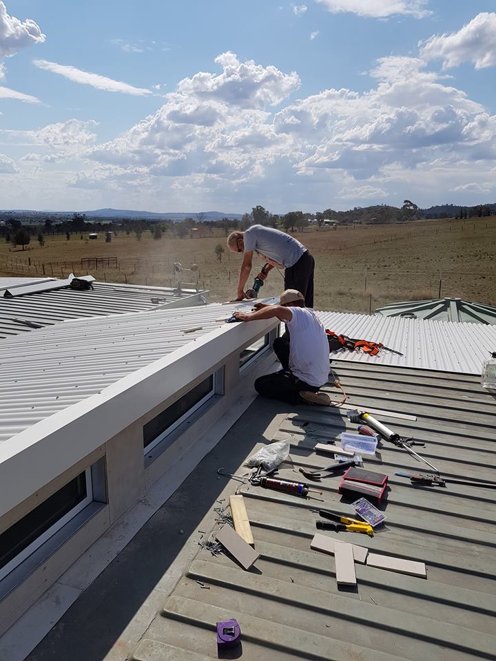Two people working on a metal roof under a partly cloudy sky. One uses a saw; the other works with tools.