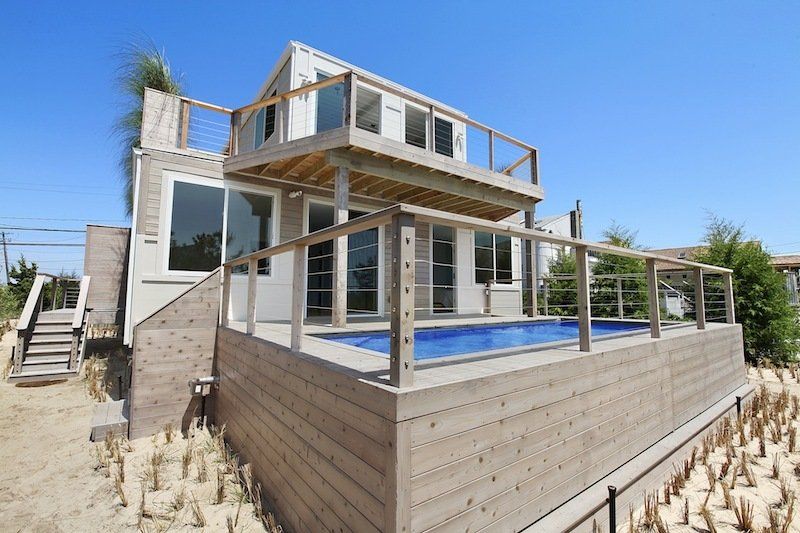 Two-story beach house with a small pool. Light wood siding and multiple decks. Blue sky and sand.