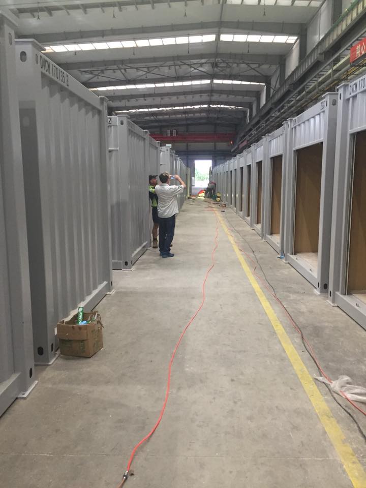 Factory interior with several gray metal containers in a row. Two people stand near the middle.