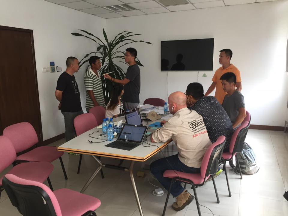 Group of men in a brightly lit room with a table, laptops, and a television, appearing to collaborate.