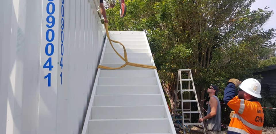 Workers attach stairs to a white shipping container. A yellow rope secures the stairs.