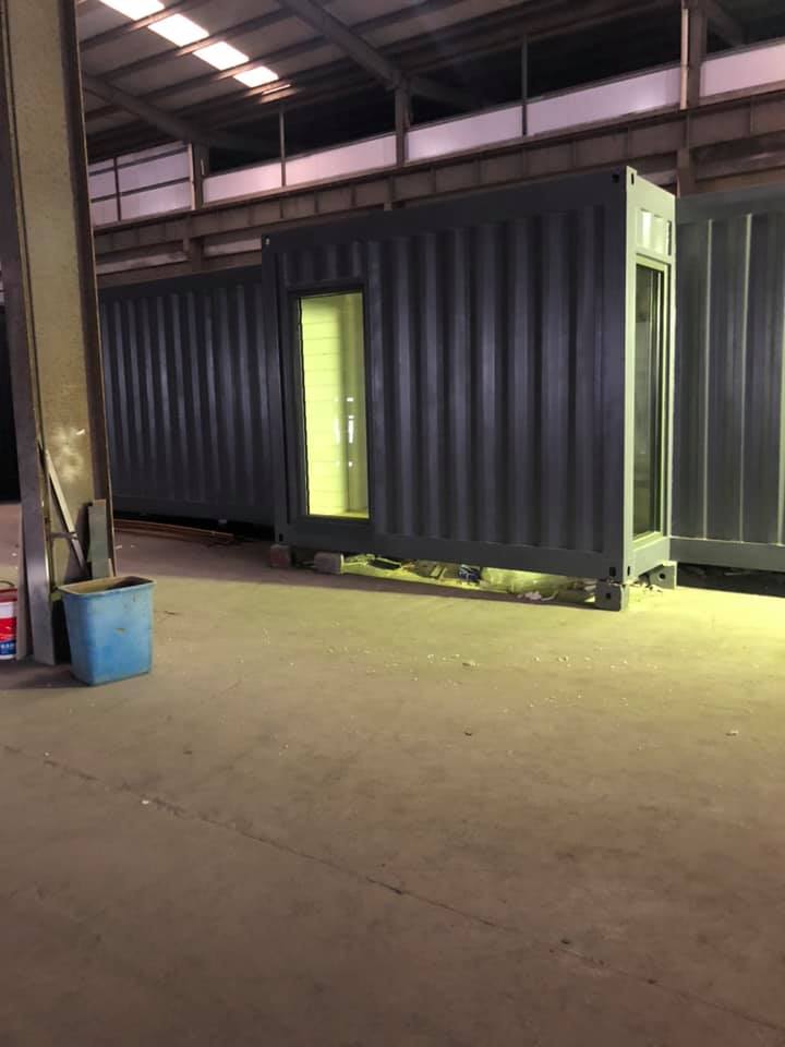 Three gray shipping containers, one with a lit doorway, inside a large warehouse.