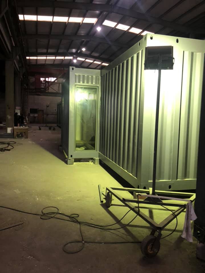 Two industrial containers inside a warehouse, one open to reveal a frame. A work light is positioned in front of them.