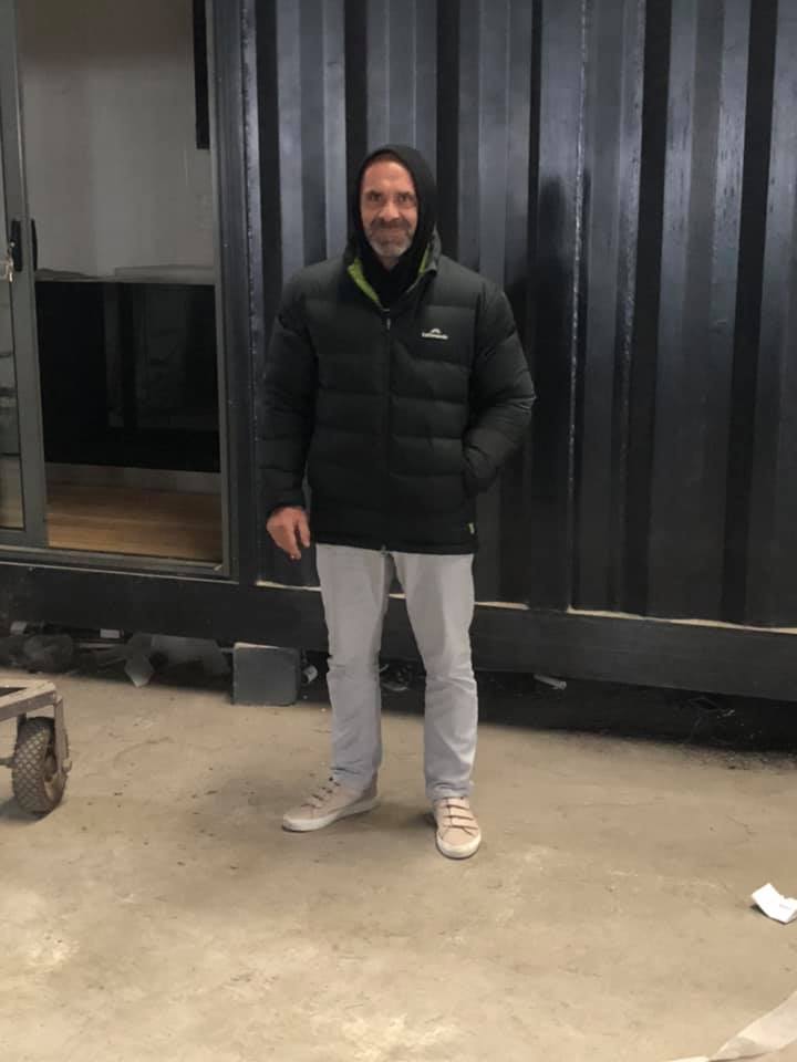 Man in black jacket and grey pants stands in front of a dark metal structure.