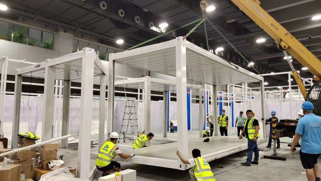 Construction workers assemble prefabricated white structures inside a large warehouse; some wear safety vests.