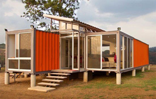 Modern orange container home with glass walls, stairs, and raised on concrete supports.
