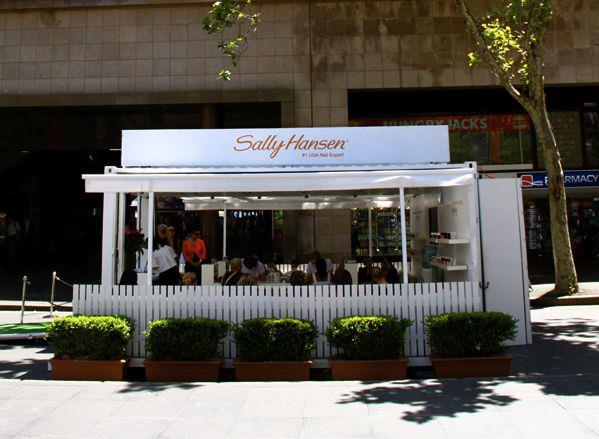 Sally Hansen pop-up store with white walls, sign, and fence. People inside, potted greenery in front. Outdoor setting.
