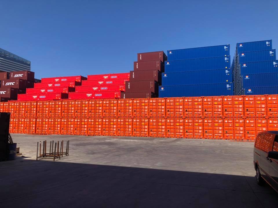 Shipping containers stacked in rows, orange, red, and blue, at a port under a clear blue sky.