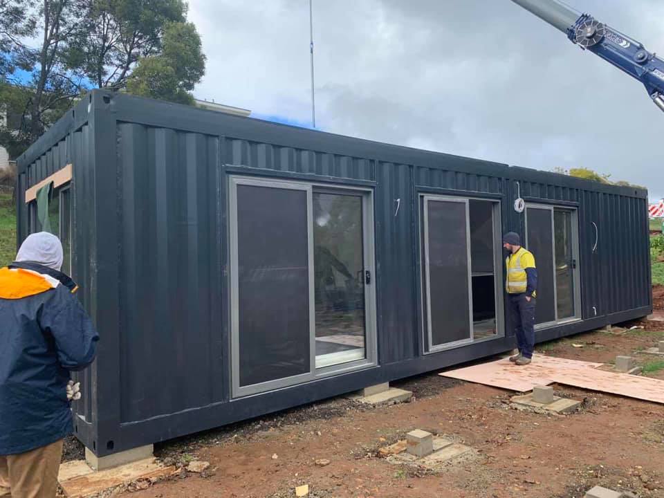 A dark shipping container, converted into a building, has sliding glass doors. Two people are near the building.