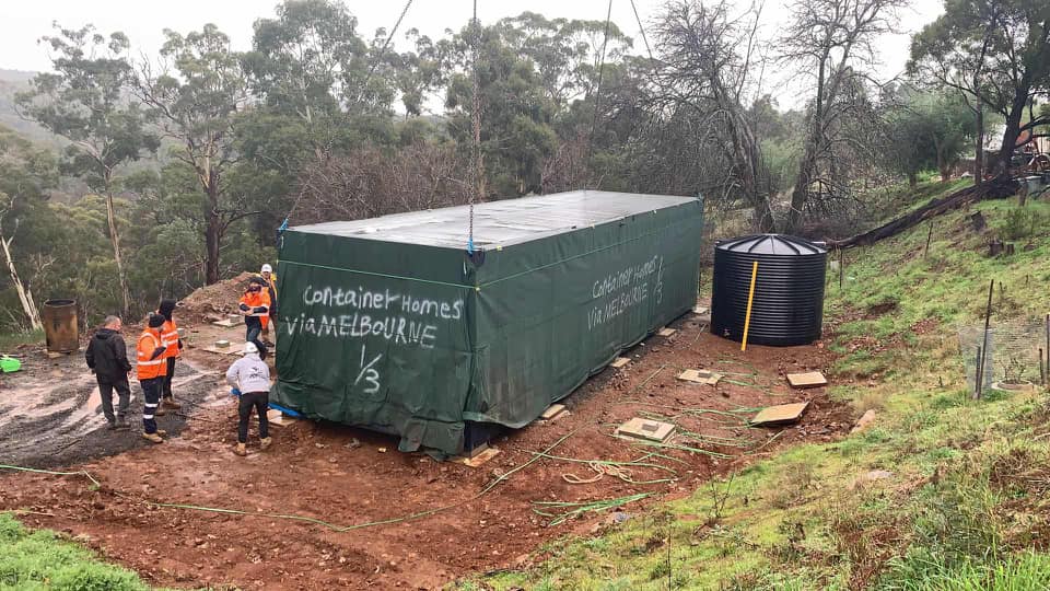 Construction site: Green container being placed near a water tank and workers on muddy ground, trees in background.