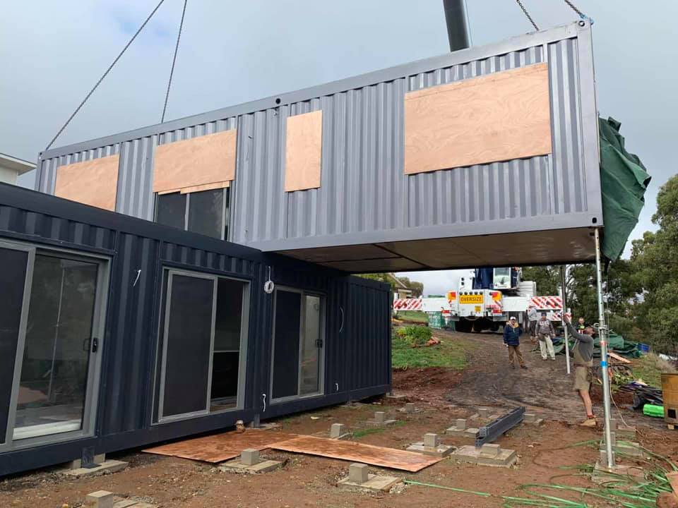 A crane lifting a modified shipping container to be joined with existing containers, construction site.