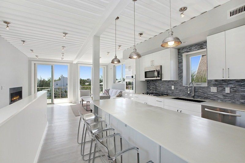 Modern, bright kitchen and living area with white surfaces, bar seating, and large windows.