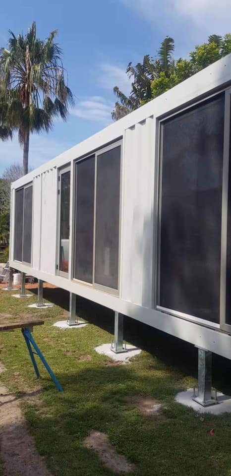 White building with large windows on metal pillars, set on grass with trees in the background.