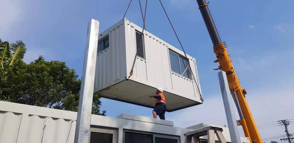 A crane lifting a white modular building with windows, placed on a white structure, worker in orange vest.