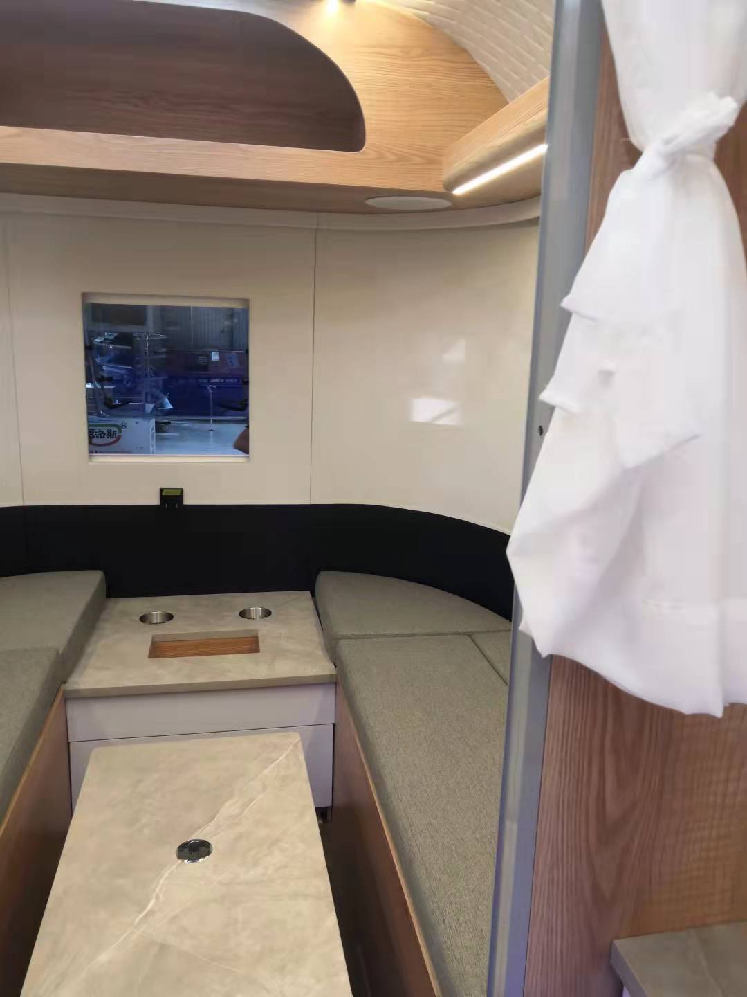 Interior of a camper, featuring seating, small window, and wooden and white paneling. A curtain is tied to the right.