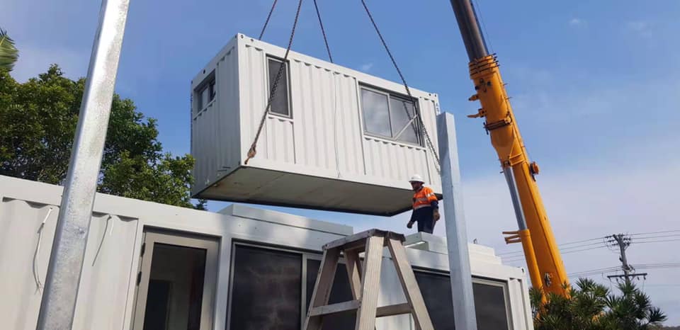 A crane lifts a white modular building component. A worker stands on a ladder, guiding the component into place on a building.