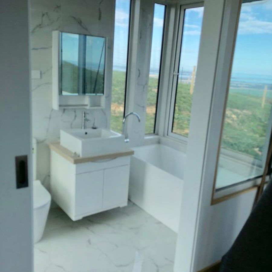 Modern white bathroom with large window, featuring sink, vanity, toilet, and bathtub.