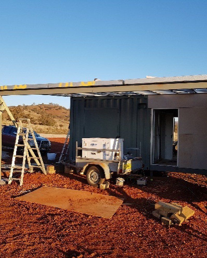 Construction site: A metal structure with a trailer, ladder, and building materials on reddish-brown dirt under a clear blue sky.