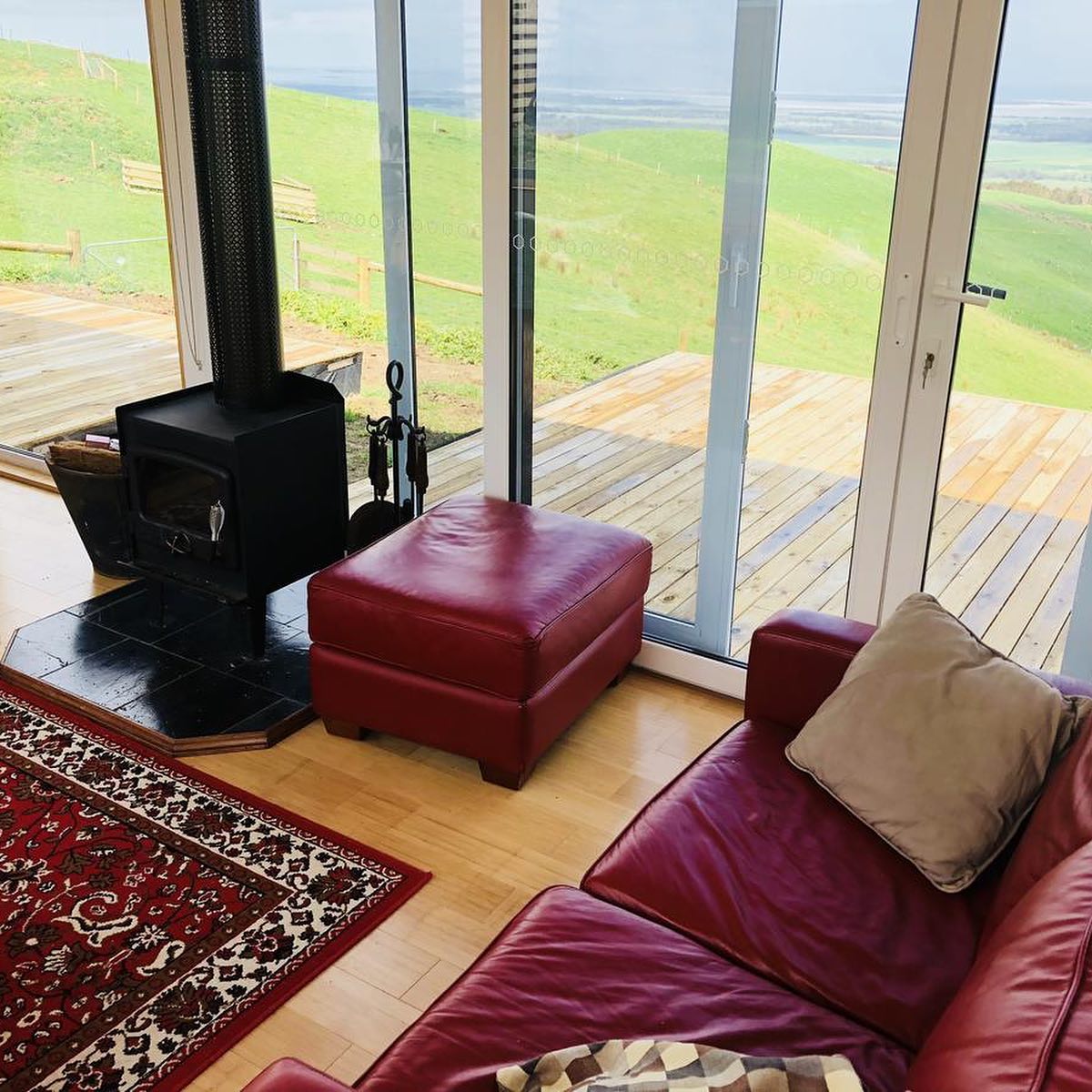 Cozy living area with red leather furniture, wood-burning stove, and panoramic window overlooking a green landscape.
