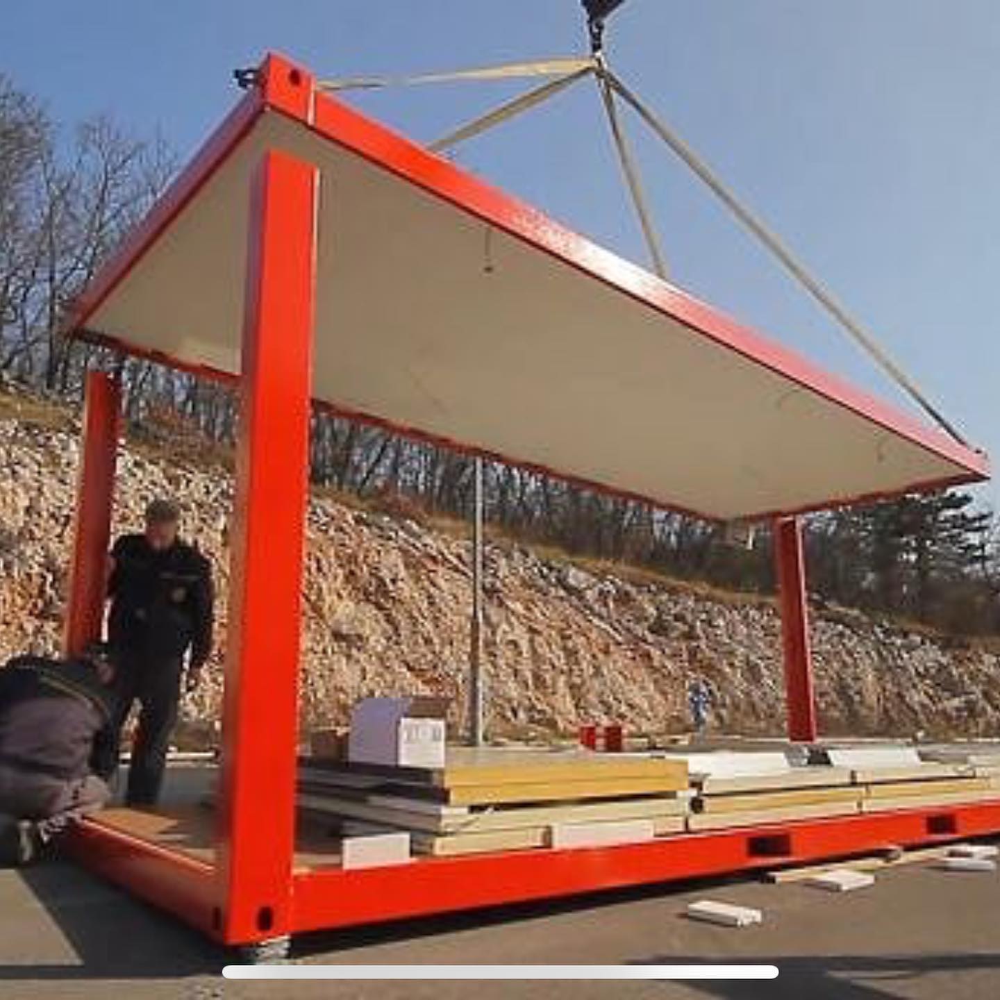 Workers assembling a red container-like structure outdoors; a crane lifts the roof into place.