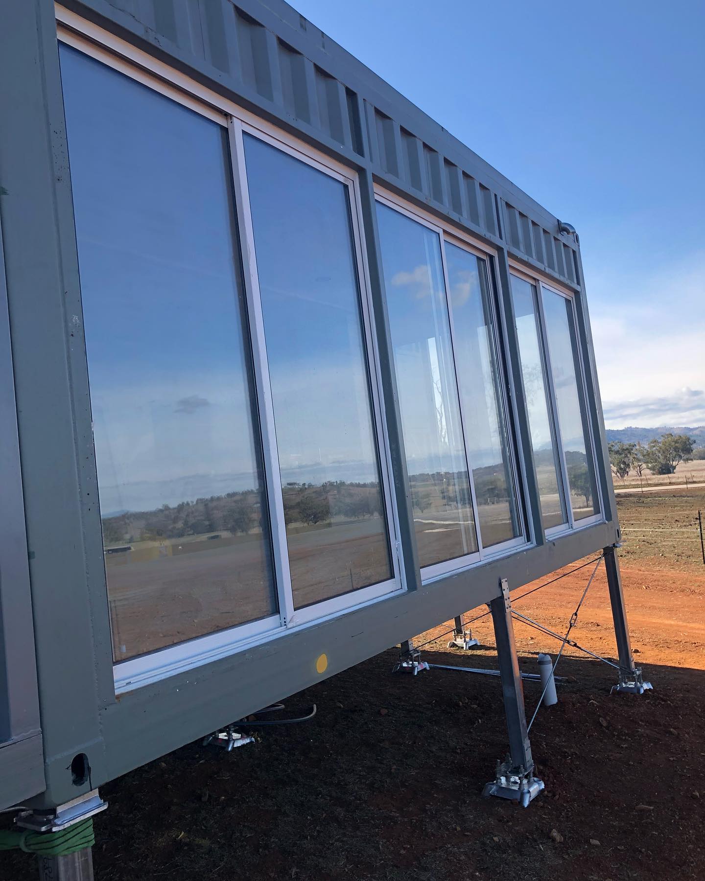 Gray shipping container home with large glass windows, elevated on adjustable legs, set against a blue sky with a scenic view.