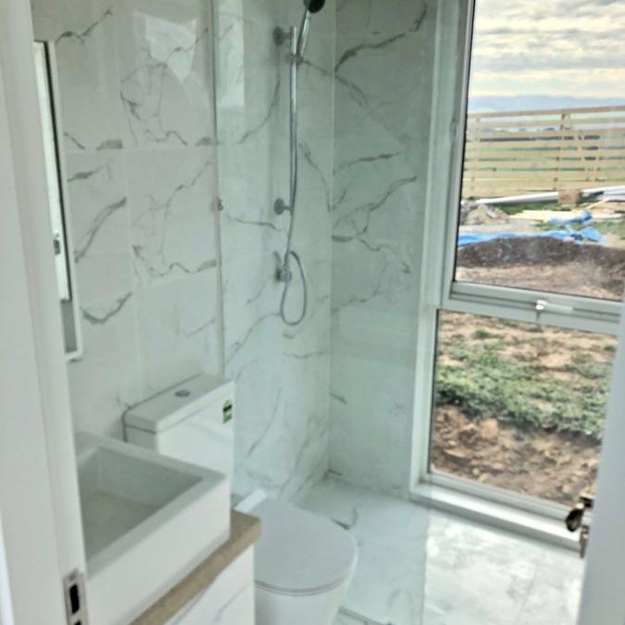 White and gray marble-tiled bathroom with a toilet, sink, shower, and a window overlooking a landscape.
