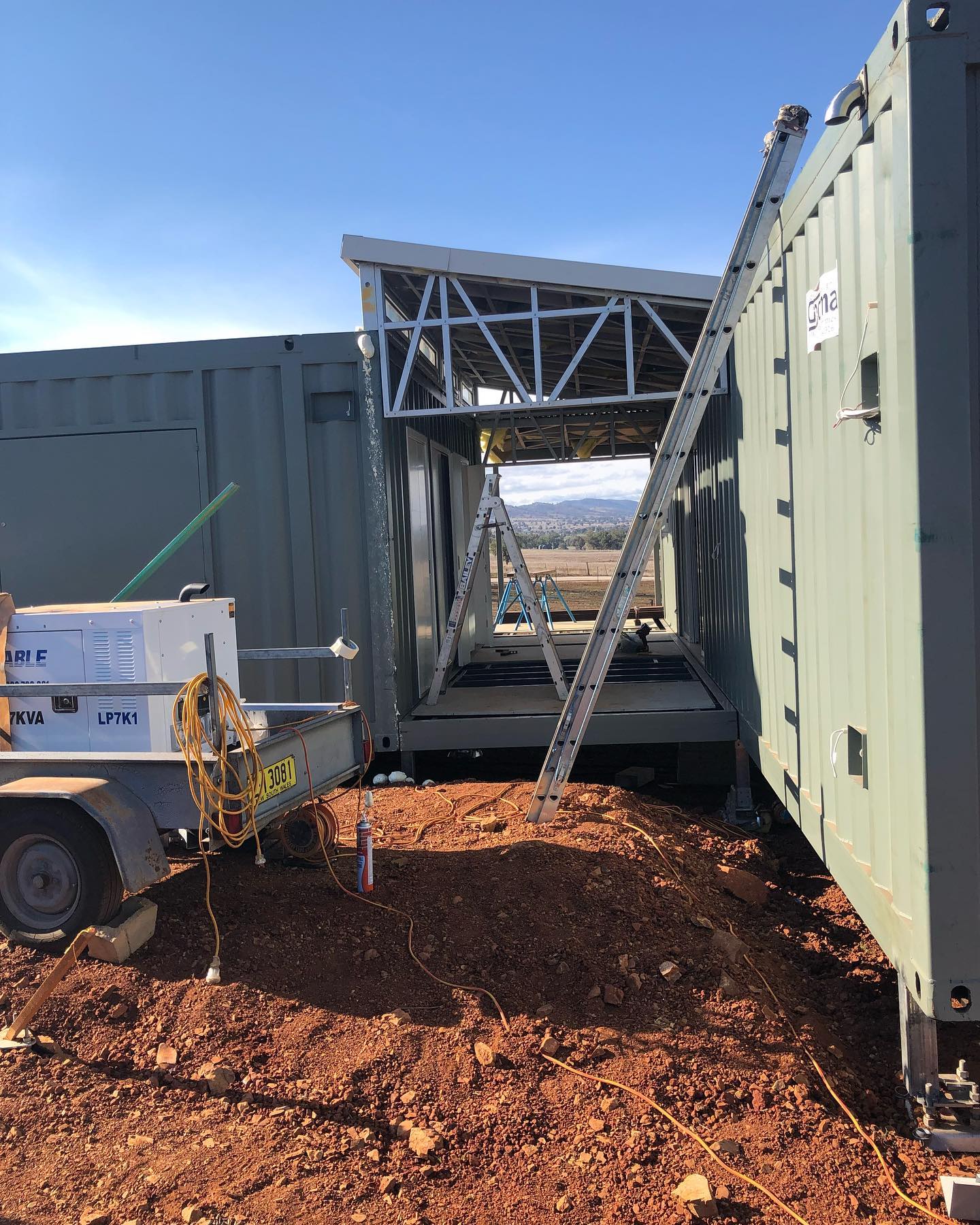 Two green shipping containers connected, with a metal frame roof. A ladder leans against one. A trailer and wires are in front.