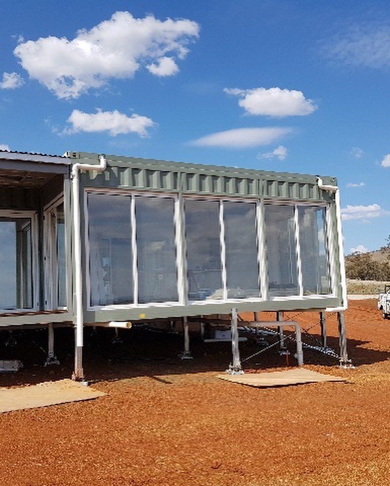 Container home with large windows, elevated on supports, set in a reddish-brown dirt area under a partly cloudy sky.