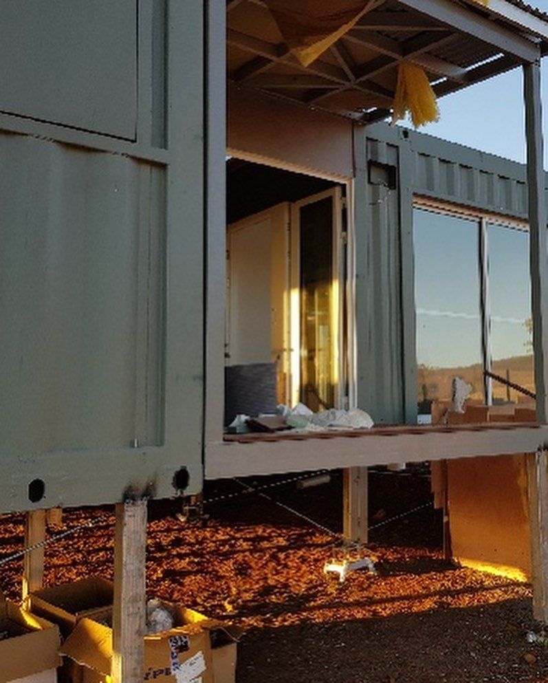 Modified shipping container home with doorway and large window, on raised supports.