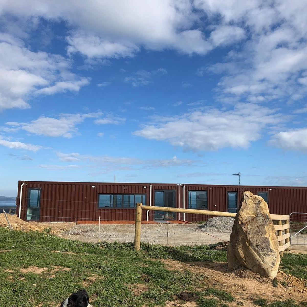 Brown shipping container home with blue windows on a hillside, fence, rock, and sky.