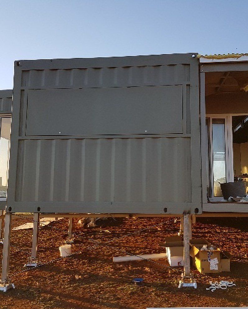Gray shipping container structure on stilts, outdoors, with a window and construction materials.
