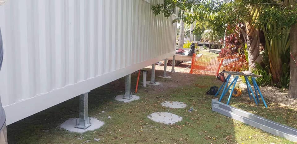A white container building elevated on concrete piers, in a grassy area with trees and construction materials.