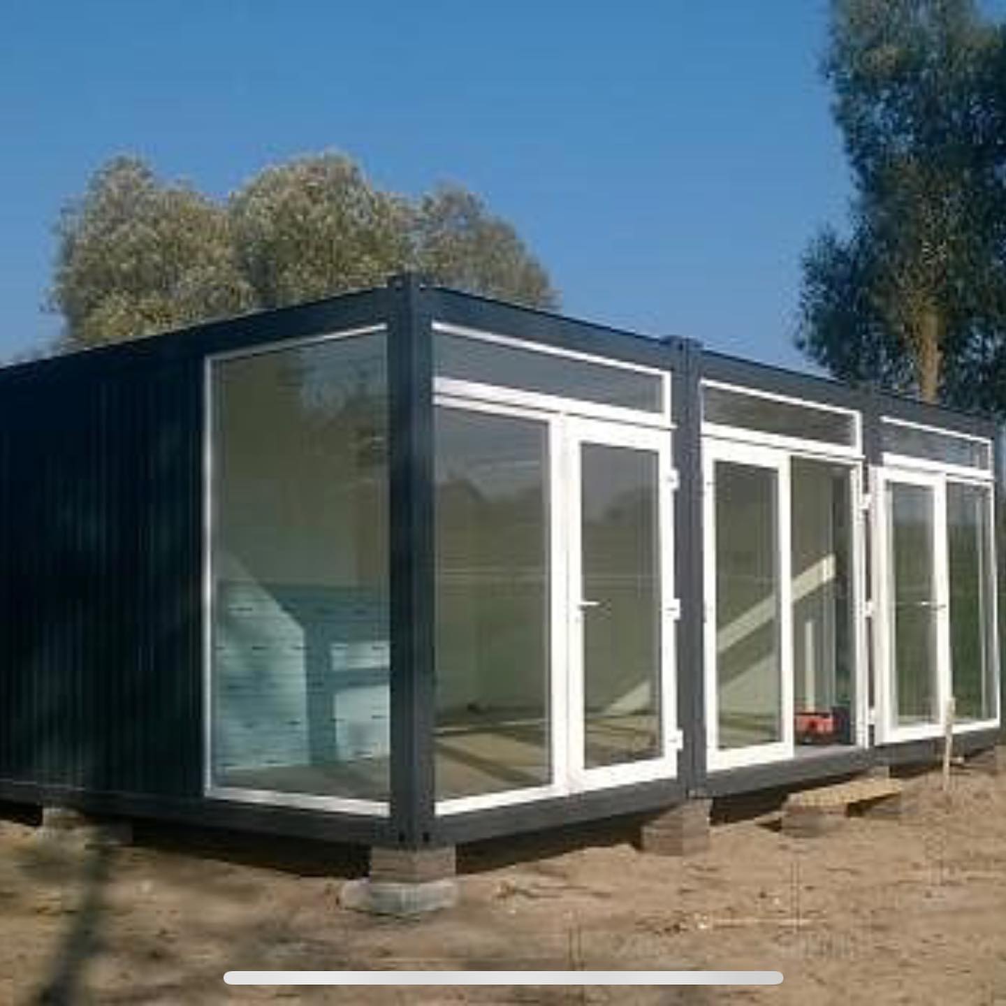 Black container-style building with large glass windows and white frames, sitting on concrete blocks outdoors.