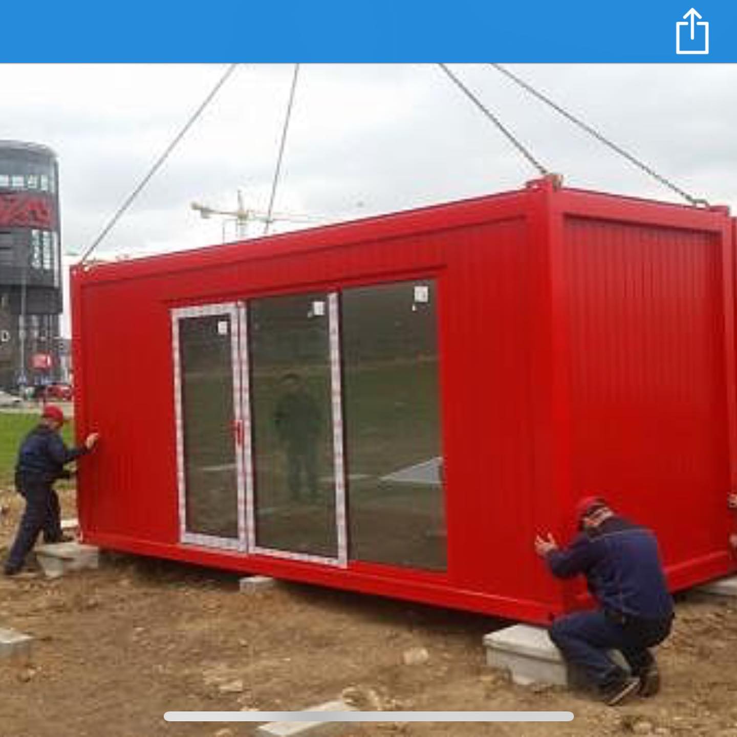 Red shipping container being lowered by crane, two workers steady it on a construction site.