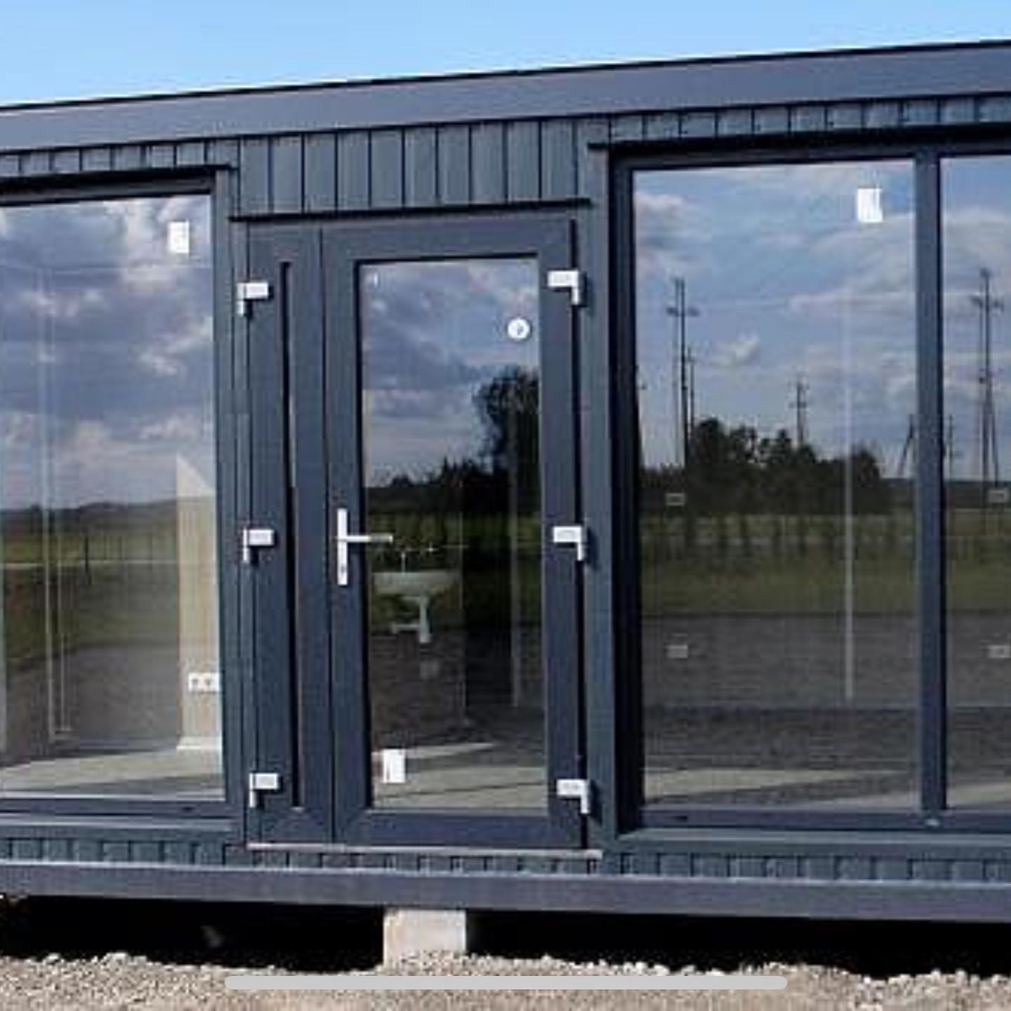 Dark gray modular building with glass doors and windows, reflecting sky and landscape.