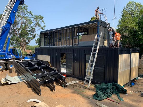 Construction of a two-story container home; workers on roof; crane nearby. Black containers with metal framing.