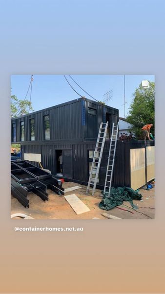 Two-story black shipping container home under construction; ladders, construction workers, and building materials present.