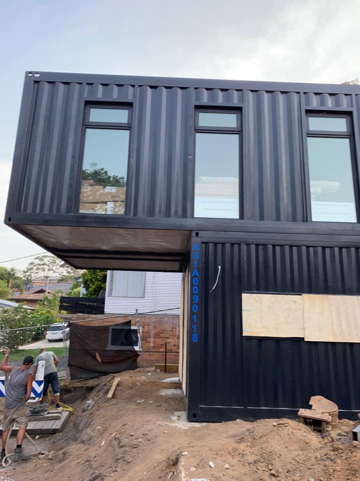 Two-story container home under construction; black containers, workers, windows.