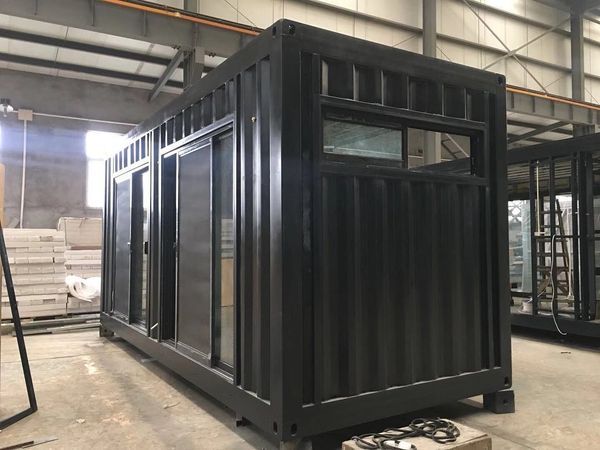 Black shipping container repurposed as a building with large windows and a sliding door, in a warehouse.