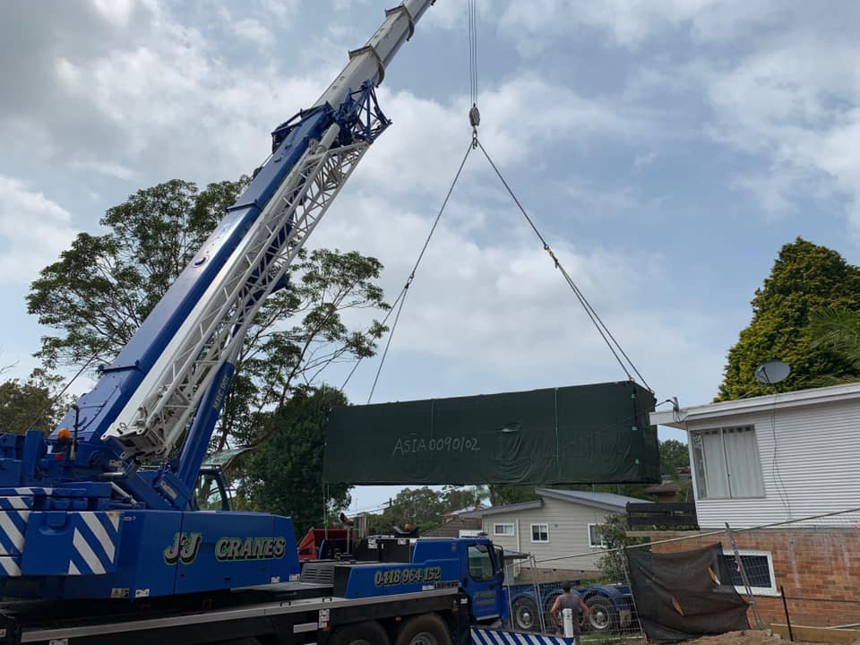 A crane lifting a large, rectangular object over residential buildings, under a cloudy sky.