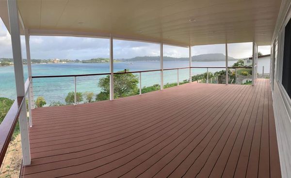 Covered deck overlooking a turquoise body of water, trees, and distant land; the deck has brown composite flooring and glass railing.