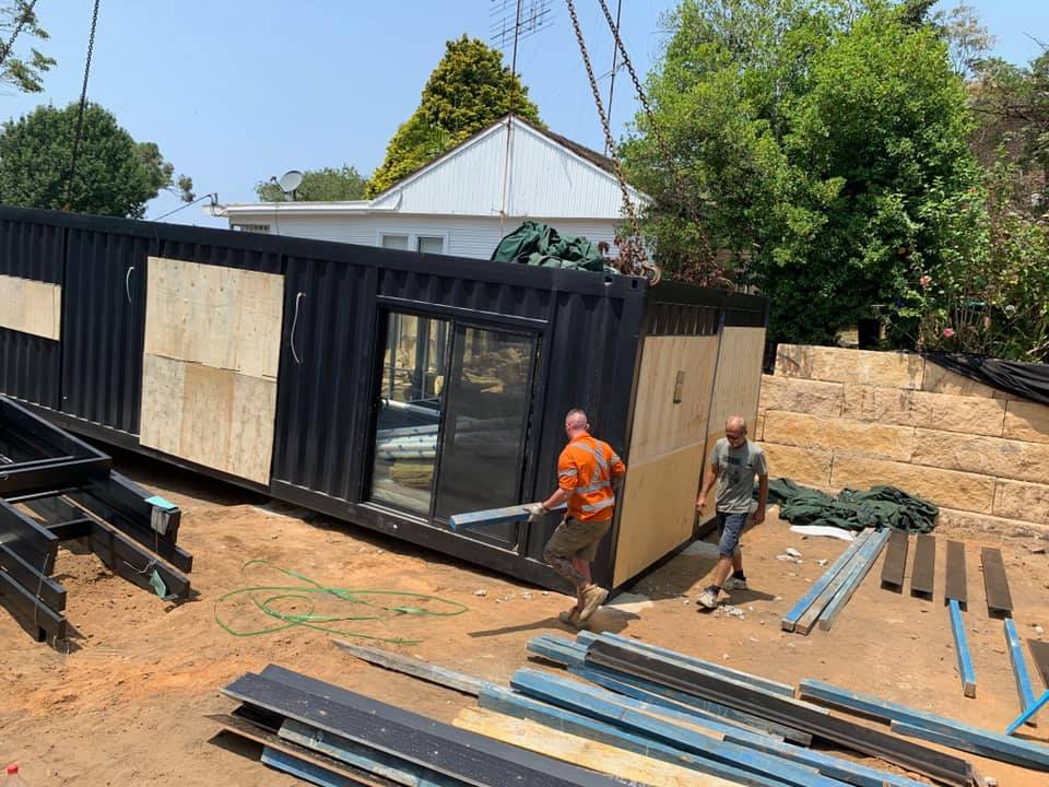 A black shipping container being installed by construction workers near a house; wooden panels and building materials on ground.