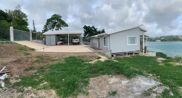 Gray house and carport beside water, with a gravel driveway and green grass.