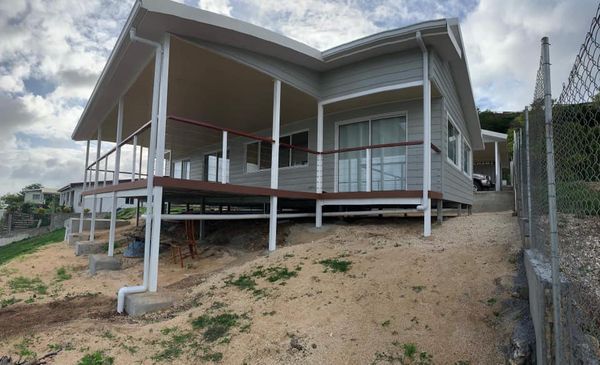 Grey house with a wrap-around porch, on a sandy hillside. Metal railing and gutter. Fence on the right.